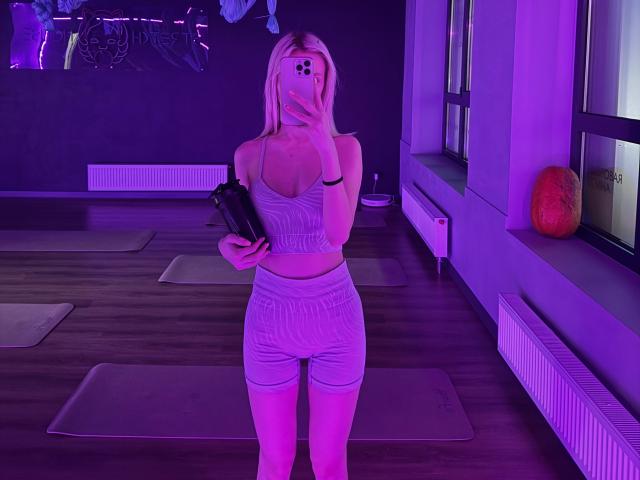 LexyGlam with blonde hair, holding a black camera, wearing a grey sports bra and matching skirt, taking a mirror selfie in a purple-lit room, showing her tits and ass.