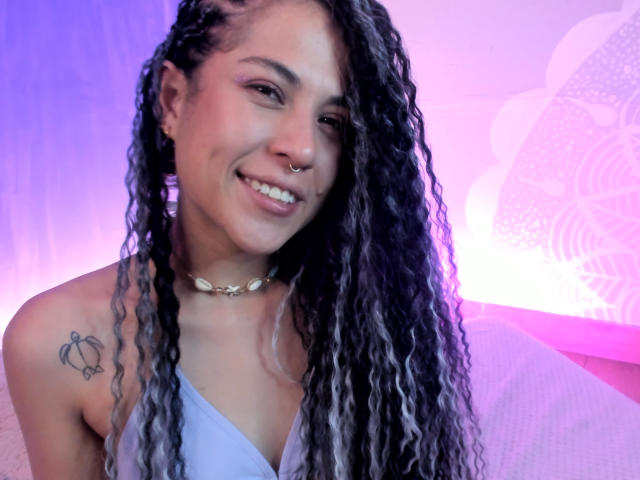 HellenK con lunghi capelli neri e bianchi ondulati, un anello al naso, che sorride, indossa un vestito blu, una collana con conchiglie e un tatuaggio di una tartaruga sulla spalla sinistra, illuminata da una luce rosa e viola..