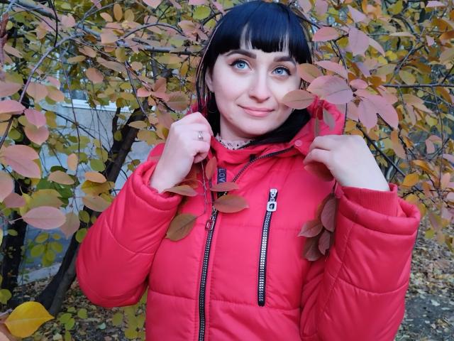 NinaFlawlees with black bangs, blue eyes, and pink lips, wearing a red puffy jacket, holding it with both hands, surrounded by yellow and brown leaves.