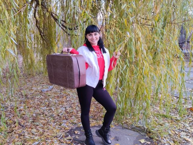 NinaFlawlees stands with a brown suitcase in her right hand, wearing a white jacket, red shirt, black pants, and black boots, under a weeping willow tree.