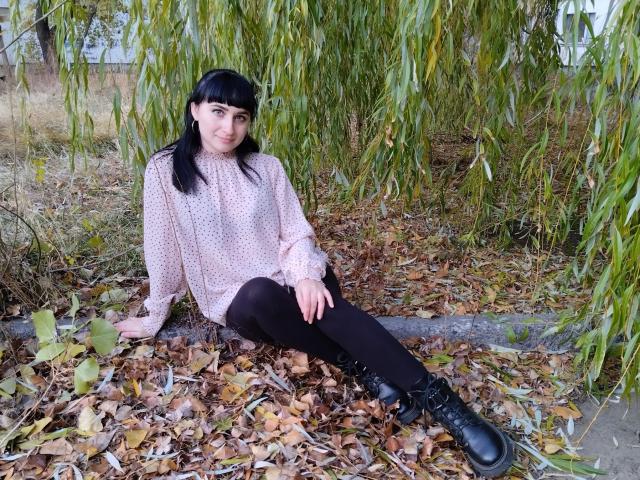 NinaFlawlees with black hair, pink polka-dot sweater, black leggings, and black boots, sitting on autumn leaves under a weeping willow, smiling at the camera.