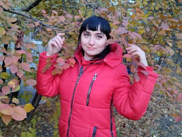 NinaFlawlees with black bangs, blue eyes, wearing a red puffy jacket, holding leaves, autumn tree in background, slightly smiling, small nose ring, hands near head.