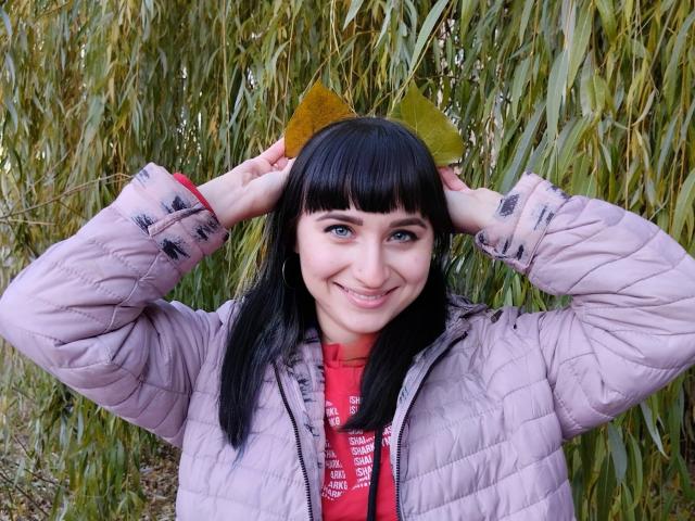 NinaFlawlees, smiling, black-haired, bangs, green olive hat, pink puffy jacket, red shirt, hands in hat, blue eyes, standing in front of tall green reeds.