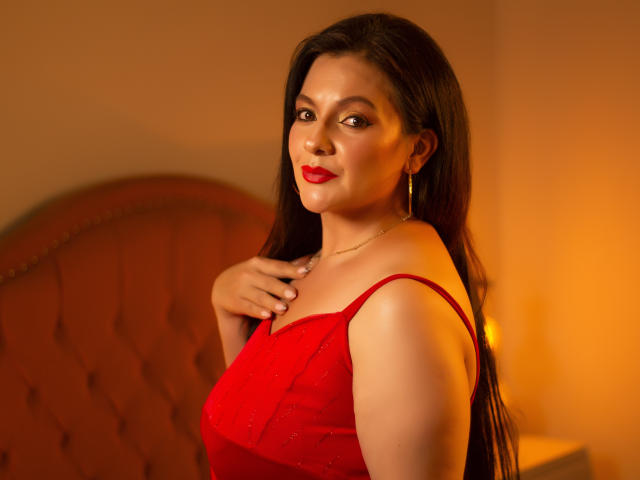 JakyKidman, a sexy woman with long black hair, red lipstick, and hoop earrings, wears a red dress, hand on chest, smiling, warm orange light, visible boobs.