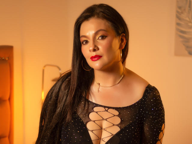 JakyKidman with long black hair, red lipstick, hoop earrings, and a black sheer dress with lace-up cleavage, standing in a warmly lit room, looking slightly to the side.