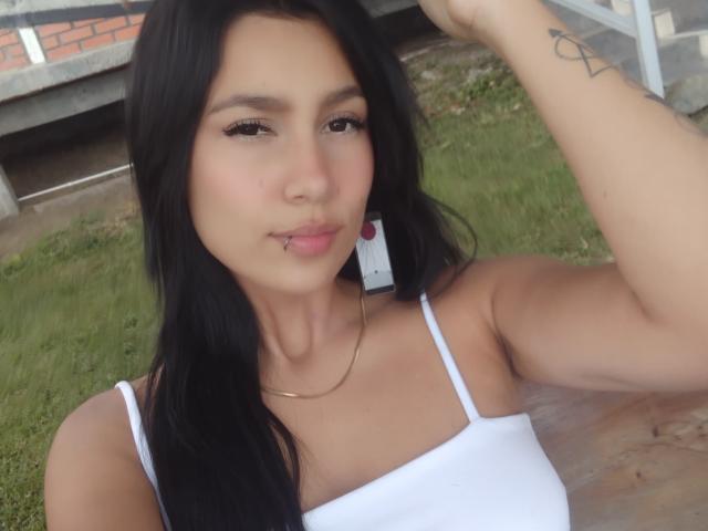 KataleyaMansson with black hair, pouty lips, wearing a white top, gold necklace, and earrings, hand in hair, tattoo on arm, looking at camera, grass in background.