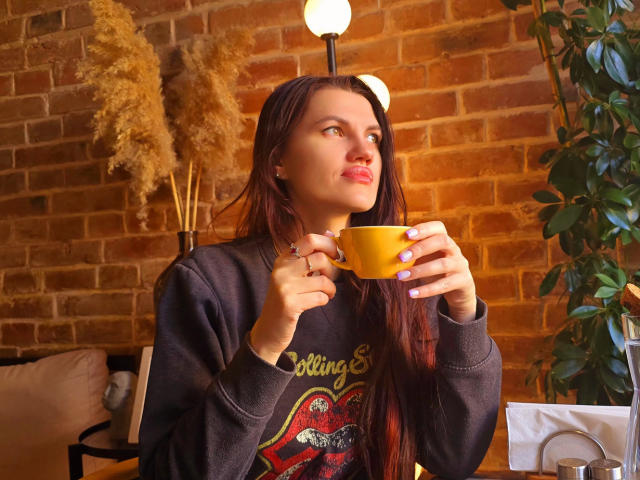 MilaLoona with long brown hair, wearing a gray sweater and gray pants, holding a large fluffy boomerang-style feather, sitting on a floral-patterned chair.