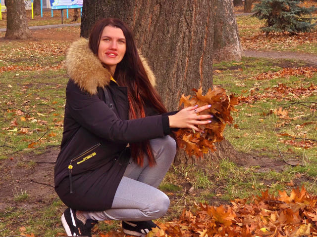 MilaLoona, squatting, holding autumn leaves, in a black coat with a fur collar, gray pants, and black shoes, has long brown hair and a cute smile, outdoors.