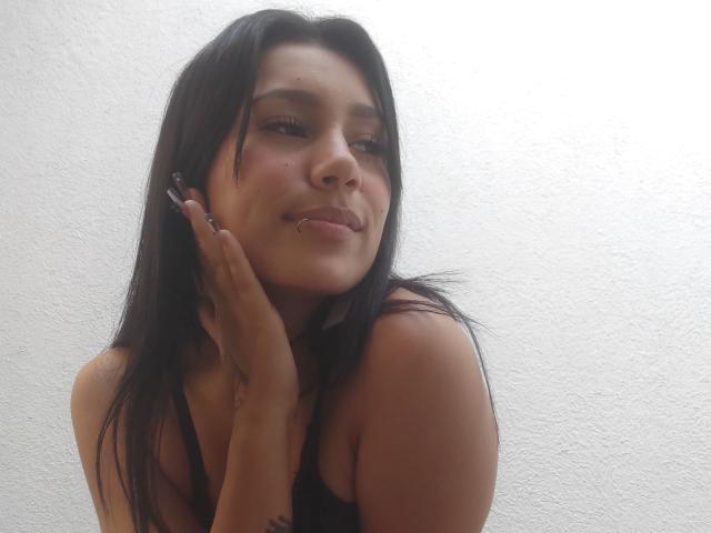 KataleyaMansson with black hair, wearing black lace bra and thong, standing against white wall, left hand on wall, right hand on belly, looking down, slight smile.