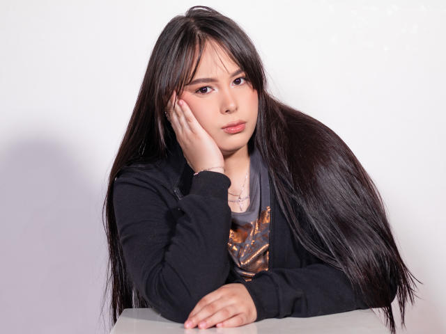BrianaRogers is kneeling on a red carpet, wearing a white off-shoulder top, exposing her large tits, with one hand covering her right nipple. She has long black hair.