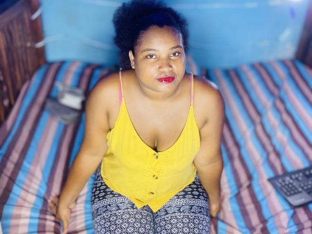 MeraBoobs with dark skin and curly hair, wearing a yellow button-up top, showing small tits, and pink bra straps, sitting on a striped bed with patterned pants.