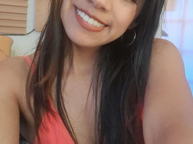 EimyRousse with long brown hair, smiling with white teeth, wearing a pink top, hoop earrings, and showing her tits in a selfie. Soft light, cute face, relaxed.