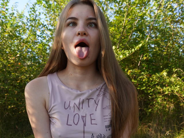EllenMays with long blonde hair, green eyes, and pink tank top, tongue out, standing outdoors, wearing "unity love" shirt, looking up with a playful expression.