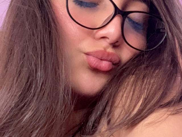 DianaFernandezz has pouty pink lips, wearing black round glasses, with long brown hair and blue eyeshadow, looking slightly to the side with a soft, cute expression.