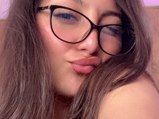 DianaFernandezz, com lábios rosados e volumosos, óculos de aro preto e cabelo castanho, piscando levemente com um sorriso divertido. Suas nádegas não estão visíveis. Seu rosto está centralizado..