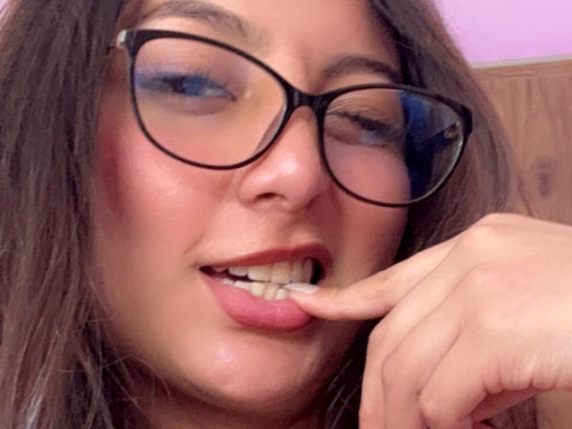 DianaFernandezz with glasses, brown hair, and pink lips, touching her lower lip with her right finger, slightly winking with blue eyeshadow visible.