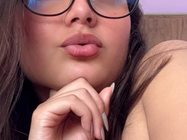 DianaFernandezz with puffy pink lips, wearing glasses, has her hand with long, white nails near her chin, brown hair, and tan skin. Boobs slightly visible, headshot.