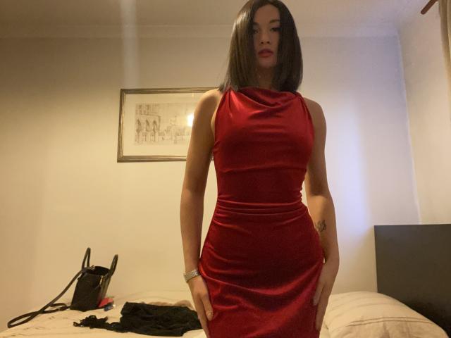 Dionisia mit glattem, schwarzem Haar, trägt ein eng anliegendes rotes Kleid, das ihre Brüste und ihren Hintern zeigt, und steht in einem Raum mit einem Bett und einer schwarzen Tasche..