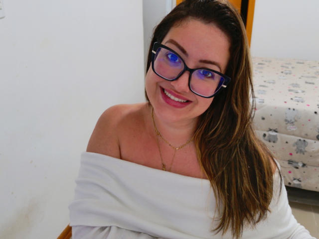 Anyellen with blue-eyed glasses, smiling, has brown hair, white off-shoulder top, and gold necklace, showing her tits. She's cute and sexy, looking directly at the camera.