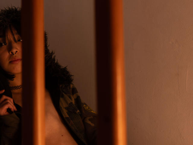 MillaHottLove69 with black curly hair, nose ring, and choker, wearing a green camo jacket with fur, partially visible through wooden bars, looking up with a slight smile.