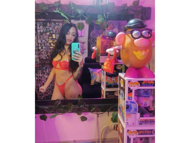 ErotiicAlexa with long black hair, red lingerie, and glasses, holding a phone, standing in front of a mirror, with a toy robot and shelves in the background.