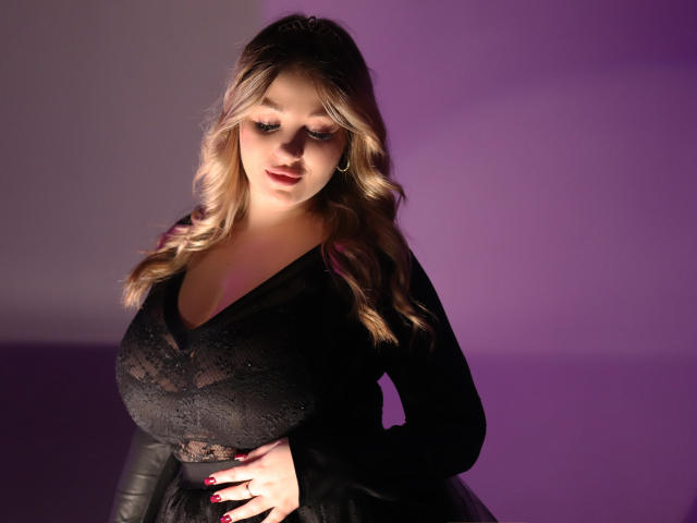 SarahStar, avec ses cheveux blonds ondulés, portant une robe noire en dentelle, montrant sa poitrine généreuse, debout devant un fond violet, une main sur la hanche, ongles rouges, lumière douce..