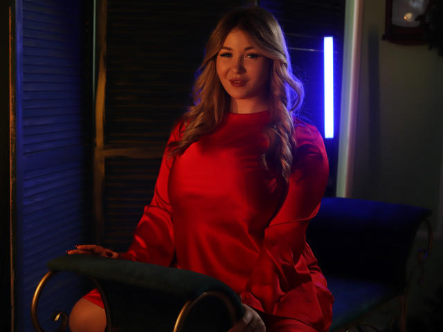 SarahStar, com longos cabelos loiros, vestindo um vestido vermelho brilhante, sentada em uma cadeira verde, com luzes azuis e roxas ao fundo, sorrindo sutilmente, com as mãos apoiadas na cadeira..