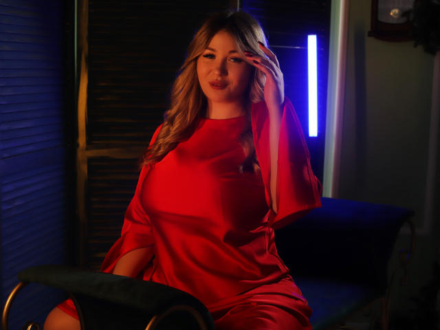 SarahStar, avec ses cheveux blonds ondulés, sa robe en satin rouge et son sourire coquin, se touche l'œil, assise sur une chaise bleu foncé. Un éclairage bleu et violet est visible en arrière-plan. Ses seins sont visibles sous la robe..
