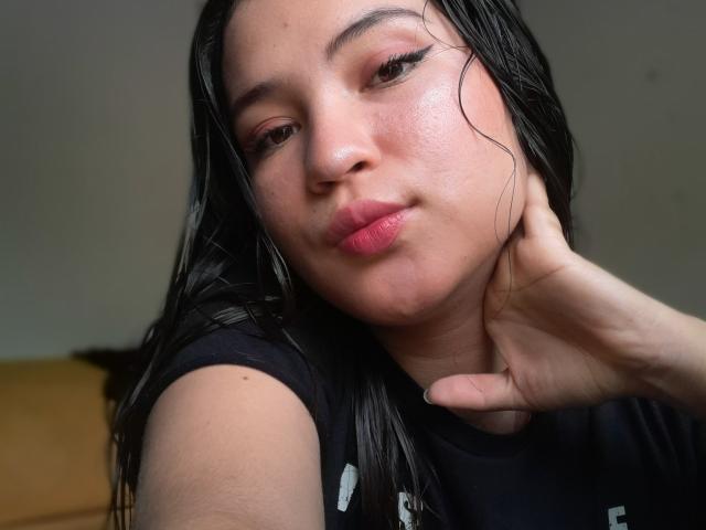 CelesteDash has wet black hair, dark eyeliner, and pink lips, pouting with a hand on her neck, wearing a black shirt, with a slightly sweaty face.
