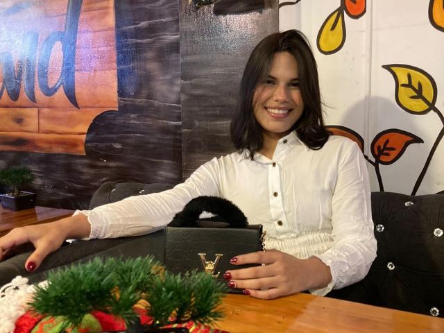 AmyXHottie, con senos de tamaño medio, vestida con una camisa blanca abotonada, sonriendo, cabello oscuro, sosteniendo un bolso negro con un borde peludo, uñas rojas, sentada en una mesa de madera..