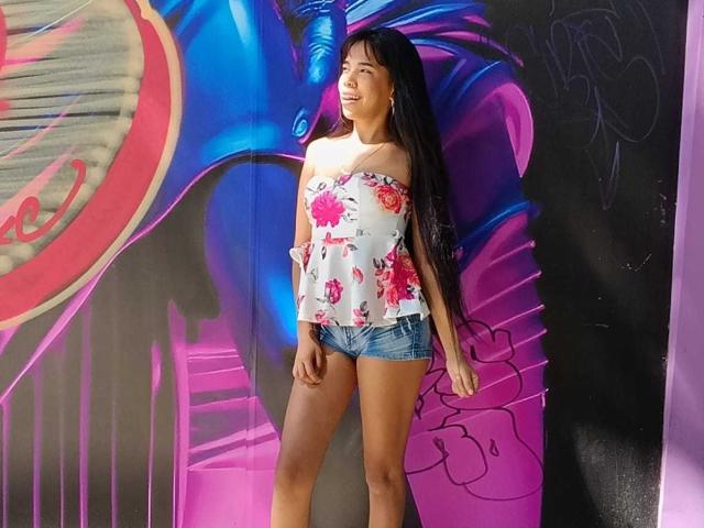 MarySmiith, com cabelos pretos longos, blusa branca com estampa floral e alças que caem do ombro, e shorts jeans, está em pé em frente a grafites coloridos, sorrindo, exibindo o bumbum e os seios de forma casual..