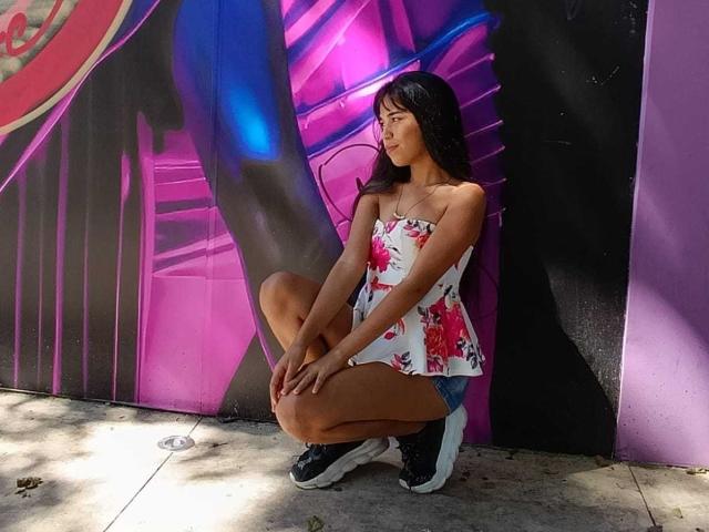 MarySmiith está en posición de sentadilla, vestida con un vestido de tirantes con estampado floral, pantalones cortos vaqueros y zapatillas negras, con cabello largo y negro, y un collar pequeño, frente a una pared de grafitis vibrante en tonos morado y rosa..