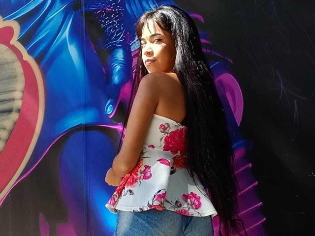 MarySmiith, con el pelo largo y negro, mirando por encima del hombro, vestida con un top blanco de flores sin tirantes y jeans azules, de pie frente a grafitis coloridos, con la luz del sol en su rostro..