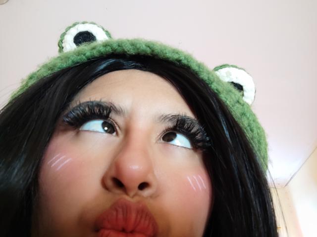 MaiaHairy's face with green frog hat, black hair, brown eyes, thick eyelashes, pink cheeks, and pouty red lips, looking up. Frog hat has green and white ears.