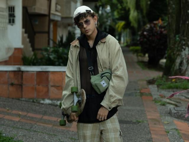AntonyGaldro, curly-haired, wearing a white cap, sunglasses, beige jacket, black hoodie, green bag, plaid pants, holding a green skateboard, standing on a brick-paved sidewalk.