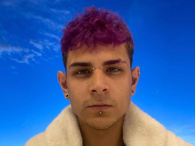 AntonyGaldro has purple curly hair, a nose ring, and a chin piercing, wearing a white furry coat, with blue sky and clouds in the background.