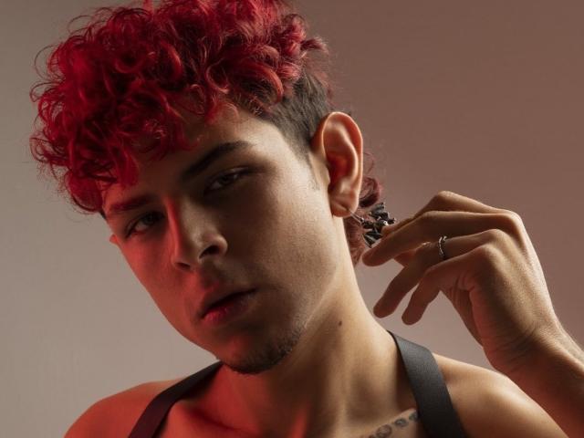 AntonyGaldro with red curly hair, dark tank top, ring on right hand, slight stubble, tattoo on chest, looking to the side, red lighting on face.