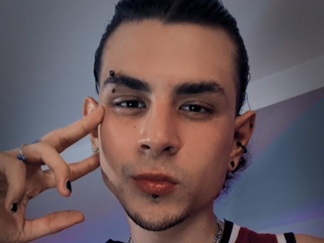 AntonyGaldro with black nose ring, black eyebrow piercing, black lip ring, black ear studs, dark hair in a bun, touching cheek with black-nailed finger, wearing red and white top.