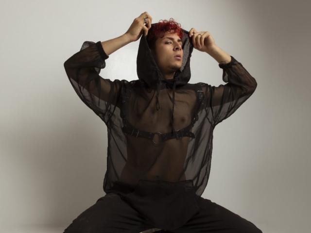 AntonyGaldro with red curly hair, wearing a sheer black hooded top revealing small tits and a black harness, pulling up the hood, sitting with legs apart, wearing black pants.