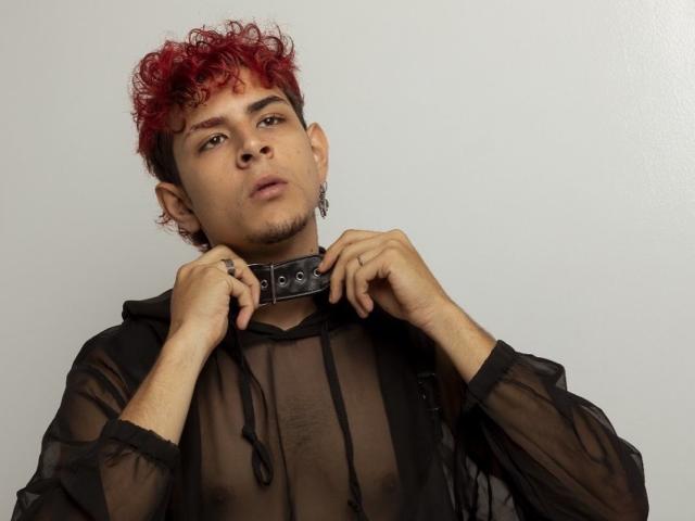 AntonyGaldro with red curly hair, brown skin, and a black mesh hoodie, holding a studded leather collar, looking up with a slight pout, wearing earrings.