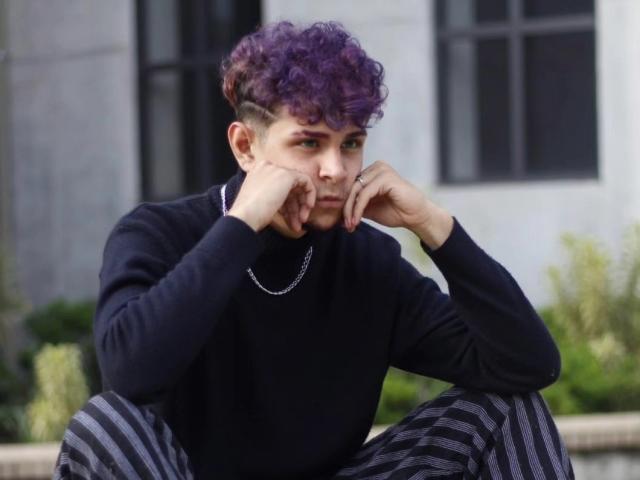 AntonyGaldro has purple curly hair, a black turtleneck, and striped pants, sitting with hands on cheeks, looking pensive. He wears a silver chain, has a short undercut.