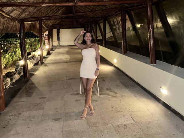 ThaliaAres stands in a wooden-covered walkway, wearing a strapless white mini-dress and sandals, her hand on her head, showing her tan skin and toned legs, with visible ass.