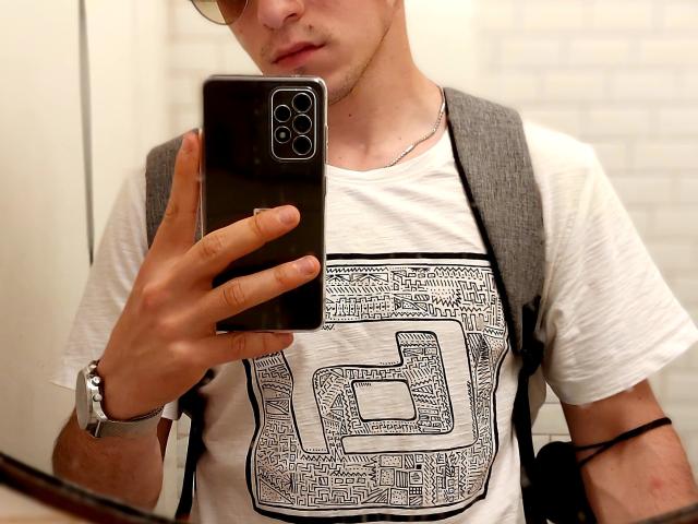 DragMan69 with glasses, holding a black iPhone, wearing a white shirt with a geometric design, gray backpack, and silver watch, taking a mirror selfie.