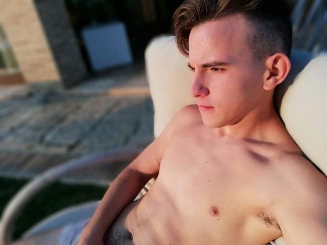 Shirtless DragMan69 with a lean, muscular build, short brown hair, and a slight tan, lying on a white chair, looking to the side with a serious expression.