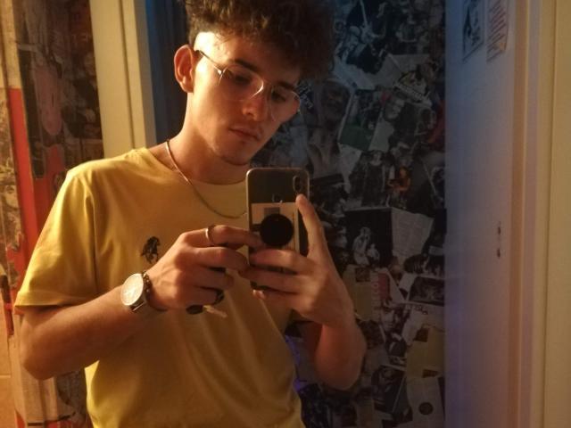 DragMan69 with curly brown hair, glasses, yellow t-shirt, and silver watch, taking a selfie in a mirror, holding a phone with a pop socket.