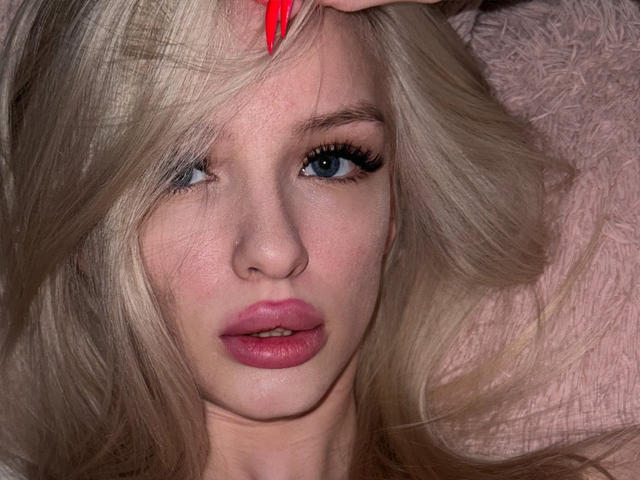 LexyGlam has blonde hair, blue eyes, and red lipstick. Her pink lips are slightly parted. She's touching her hair with red nails. Her face is flushed and close-up.