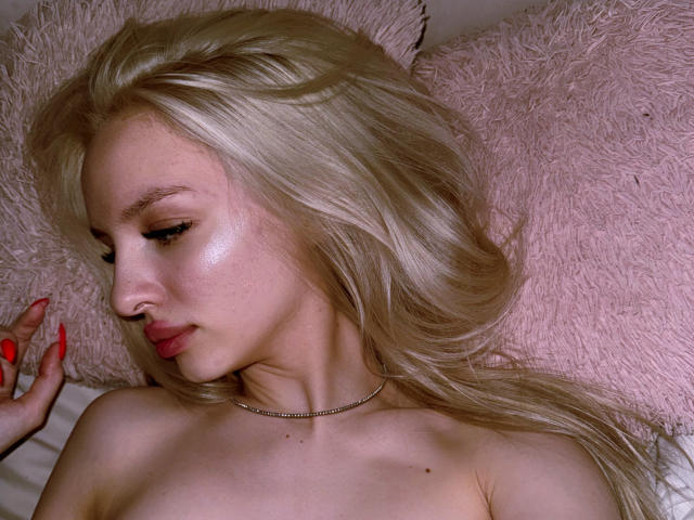 LexyGlam, blonde, topless, lying down, pink fluffy pillow, shiny skin, red nails, subtle makeup, side profile, necklace, long wavy hair, soft light, relaxed, sexy, upper body.