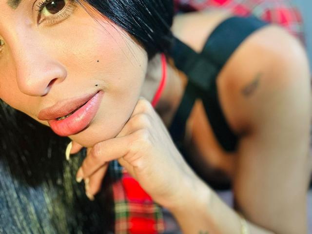 O rosto de Stefeney Collin, com cabelo preto, olhos castanhos, uma pequena marca de beleza e lábios rosados e brilhantes. Ela está vestindo uma blusa preta com um colar vermelho, e sua mão está apoiada no queixo..
