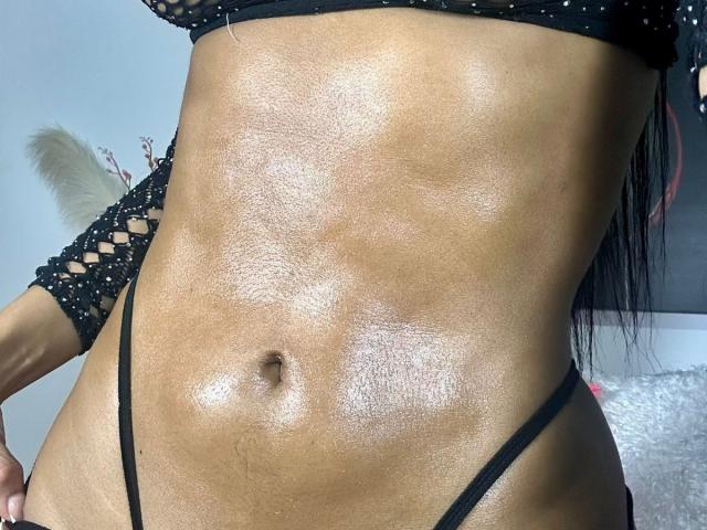 La barriga brillante y aceitada de Stefeney Collin, con una pequeña tanga negra, que muestra un abdomen tonificado y musculoso, y una camiseta de malla negra con mangas recortadas. Se puede ver el contorno de los senos..