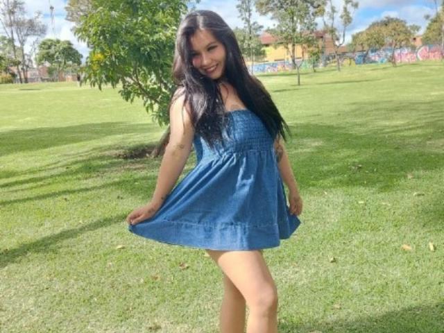 NinnaNaughty with long black hair, smiling, wearing a blue dress, holding the skirt, standing on grass, showing her bare ass, in a sunny park.